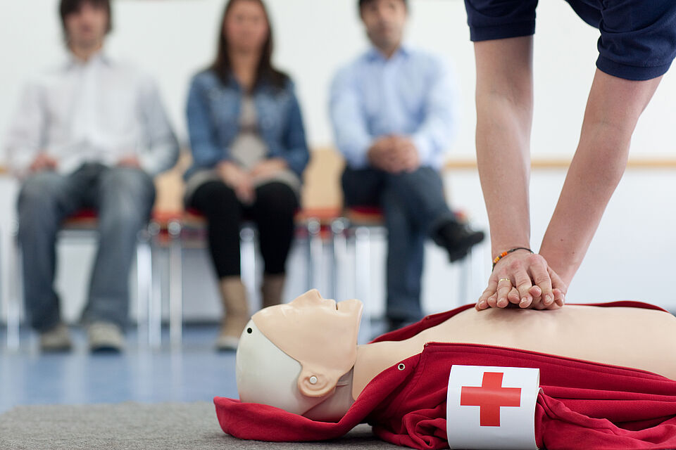 3 Menschen im Hintergrund im Vordergrund eine Puppe und eine simulierte Wiederbelebung mit 2 Händen auf der Brust 